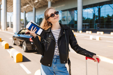 Fototapeta premium Beautiful smiling girl in sunglasses and leather jacket with passport and flight ticket in hand while happily looking in camera near airport with cabriolet car on background