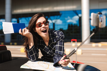 Joyful girl in sunglasses with map leaning on cabriolet car while happily taking photos on...