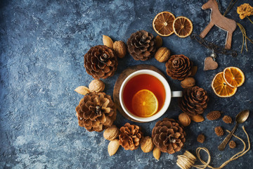 Obraz na płótnie Canvas Cozy autumn morning with cup of tea with lemon, decorative corns, dried oranges, nuts and wooden toys on dark blue background, teatime, hugge concept, selective focus, top view