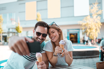 Young man with sunglasses is taking selfie with his girlfriend while they having ice-cream outdoors