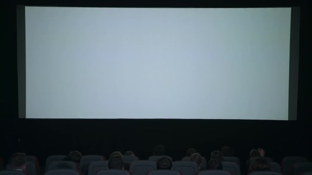 Spectators applauded in anticipation of movie session sitting in front of large white screen. White screen in cinema in slow motion. Spectators looking on cinema white screen. Movie entertainment