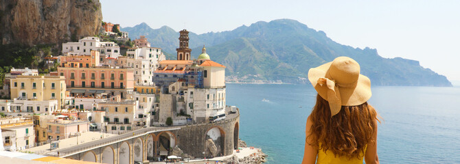 Summer holiday in Italy panorama banner. Back view of young woman with straw hat and yellow dress with Atrani village on the background, Amalfi Coast, Italy.