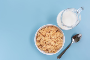 top view of corn flakes in a bowl with jug of milk and stainless spoon on light blue background. quick breakfast for modern lifestyle concept.