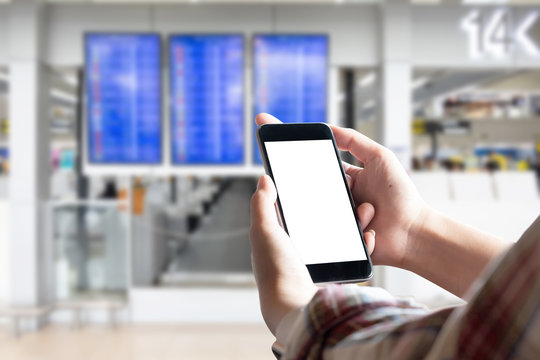 Mockup image of female hands holding black mobile phone with blank white screen over flight board in airport terminal.