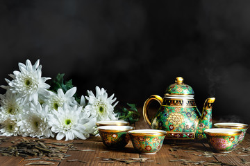 Still life of porcelain tea set of pot and cups with white chrysanthemum flowers on wooden table with scattered dry tea leaves on dark and smoky background