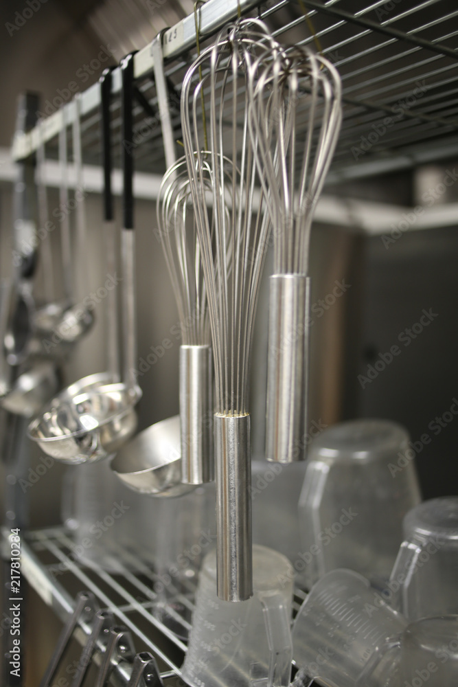 Wall mural wire whisks hanging from a stainless steel silver metal rack in an industrial kitchen