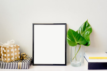 Black photo frame, green plant in a chrystal vase, gift boxes and a pile of books arranged against emty grey wall. Frame mock-up.