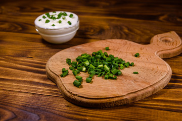 Glass bowl with sour cream and cutting board with chopped green onion on wooden table