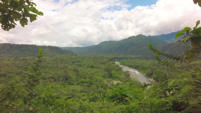 A high-angle view of the meandering Mulatos River through a dense green tropical forest valley near Tena in the Ecuadorian Amazon, foliage in the foreground and mountains under a cloudy sky.