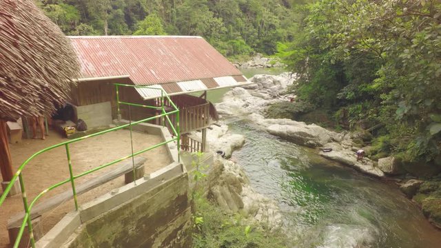 A high-angle view of a rustic thatched-roof hut beside the rocky Mulatos River at Tena Laguna Azul in the Ecuadorian Amazon, people near the water, the camera panning right.