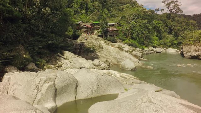 A wide shot of the green Mulatos River flowing through a lush jungle valley near Tena in the Ecuadorian Amazon, large smooth rocks and traditional wooden huts on the bank, the camera panning right.