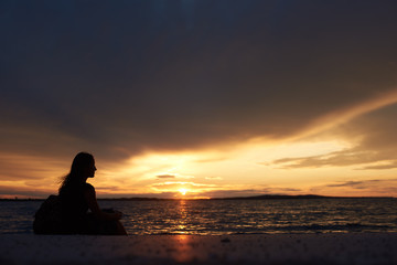 Naklejka na ściany i meble Silhouette of profile tourist woman with backpack sitting alone on seashore at water edge, enjoying beautiful view of sunset on dark evening sky background. Tourism and vacations concept.