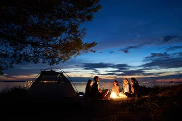 Wall murals Camping Night summer camping on lake shore. Group of five young tourists sitting on the beach around campfire near tent under beautiful blue evening sky. Tourism, friendship and beauty of nature concept.