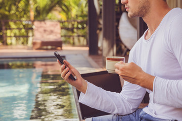 Attractive man using cellphone and drinking coffee while sitting near swimming pool.