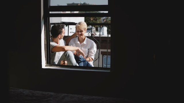 Camera approaching beautiful young romantic couple sitting close together, hugging and talking at a small sunny balcony.