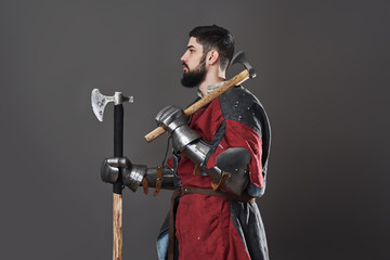 Medieval knight on grey background. Portrait of brutal dirty face warrior with chain mail armour red and black clothes and battle axe