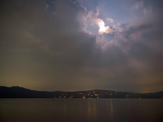 Naklejka na ściany i meble 夜の湖畔にて、空には雲が一面に広がり、隙間から月明かりが放射線状に伸びる、家明かりが灯る。