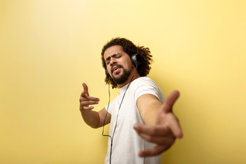 A curly-headed tanned man wearing headphones is pointing at the camera with his eyes closed.