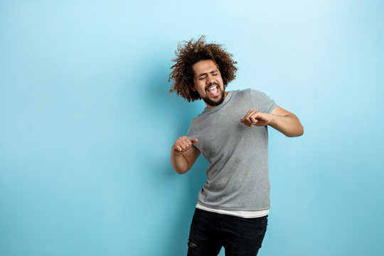 A curly-headed handsome man wearing a gray T-shirt is standing with a cheerful smile and dancing with his eyes closed over the blue background.