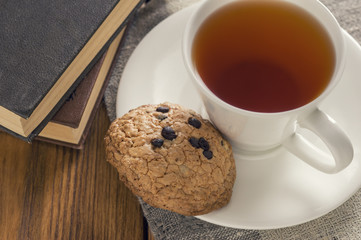 A cup of tea and some chocolate chip cookies over a books on a brown wooden table.