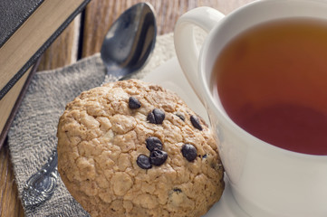 A cup of tea and some chocolate chip cookies over a books on a brown wooden table.