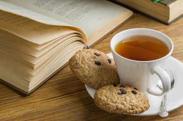 A cup of tea and some chocolate chip cookies over a books on a brown wooden table