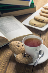 A cup of tea and some chocolate chip cookies over a books on a brown wooden table
