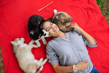 A beautiful smiling woman with a ponytail and wearing a striped shirt is cuddling with  three sweet husky puppies while resting on the red blanket on the lawn. Love and care for pets. © Leika production