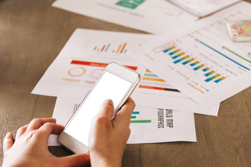 Human hands using an empty and blank phone screen with data and information analysis papers on desk.