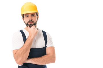 serious construction worker in hard hat standing with hand on chin and looking at camera isolated on white