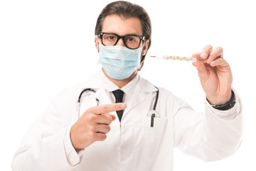 doctor in medical mask pointing at thermometer and looking at camera isolated on white