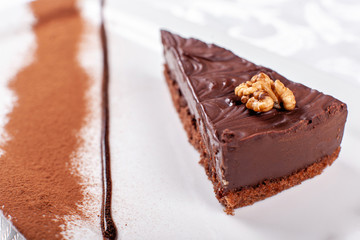 Piece of chocolate cake, cocoa biscuit with walnut filling and dark chocolate icing served on a plate.