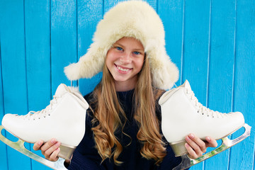 Cheerful little girl in warm sweater and hat keeps figure skates. Blue wooden background.
