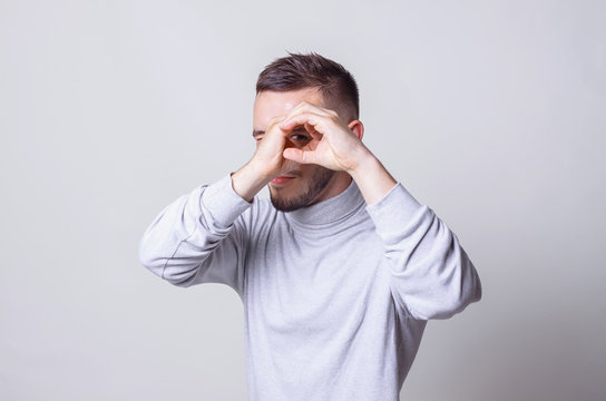 Portrait of young playful man holding hands near eyes in a shape of imaginary spyglass on grey background copy space