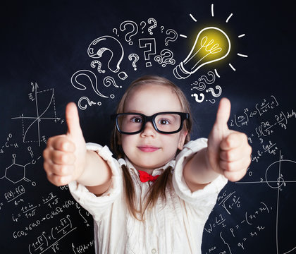 Happy pupil little girl showing thumb up in classroom on blackboard background with question mark and lightbulb
