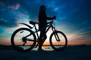 Silhouette of a young woman with a bicycle on the lake at sunset