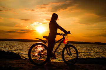 Fototapeta premium Silhouette of a young woman with a bicycle on the lake at sunset