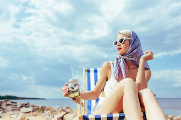 attractive fashionable girl holding lemonade and relaxing in deckchair on rocky shore