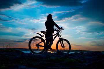 Fototapeta premium Silhouette of a young woman with a bicycle on the lake at sunset