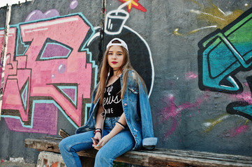 Stylish casual hipster girl in cap and jeans wear listening music from headphones of mobile phone against large graffiti wall with bomb.
