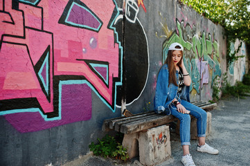 Stylish casual hipster girl in cap and jeans wear listening music from headphones of mobile phone against large graffiti wall with bomb.