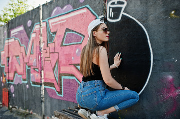 Back of buttocks stylish casual hipster girl in cap, sunglasses and jeans wear, listening music from headphones of mobile phone against large graffiti wall with large tnt bomb.