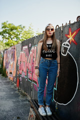 Stylish casual hipster girl in cap, sunglasses and jeans wear against large graffiti wall with large tnt bomb.