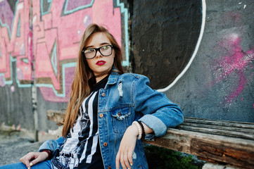 Stylish casual hipster girl in jeans wear and glasses against large graffiti wall.