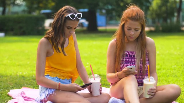 leisure, technology and internet addiction concept - happy smiling teenage girls or friends with smartphones and milk shakes or smoothie at picnic in summer park