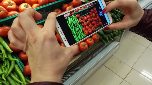 Hand taking picture of tomatoes and other vegetables using smart phone at grocery store.