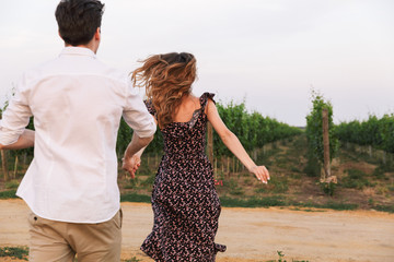 Photo from back of young caucasian couple man and woman walking outdoor together, through vineyard on summer day