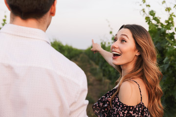 Fototapeta premium Joyful young couple man and woman smiling while walking outdoor together, through vineyard on summer day