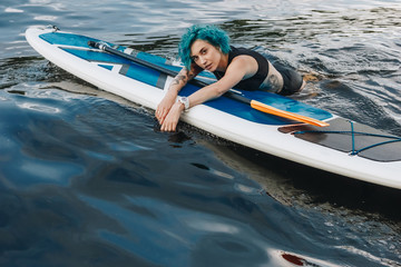 beautiful athletic tattooed young woman with blue hair relaxing on sup board in water