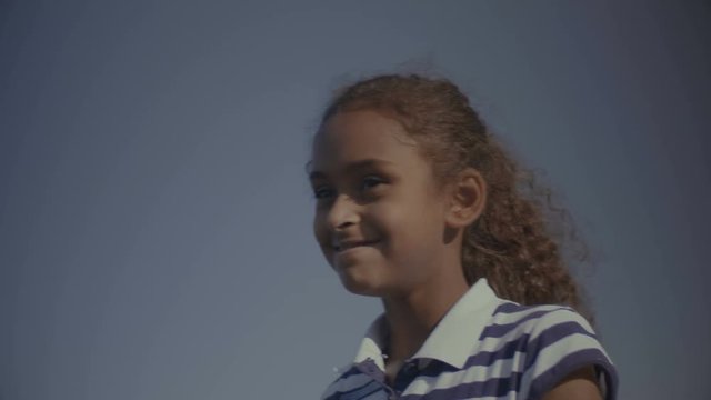 Outdoors close up portrait of smiling cute african american elementary age girl against blue sky. Carefree joyful black preteen curly child looking away with innocent toothy smile and laughing.